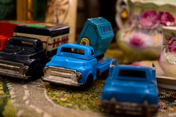 A collection of vintage toy trucks arranged on a textured surface. The bright blue and black toy trucks have a worn appearance, suggesting they are well-loved and possibly used frequently in play. In the background, there are intricately designed ceramic teacups with floral patterns, adding a nostalgic feel to the overall setting.