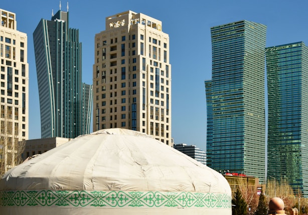 A traditional yurt with decorative patterns sits in the foreground, while modern skyscrapers rise in the background. The sky is clear and blue, providing a contrast between the traditional and the contemporary.