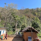 A rural setting with a thatched-roof hut and a solar panel in the foreground, surrounded by lush greenery and hills. A woman and a child are seen interacting, while another person stands near the hut, engaging in daily chores under a clear blue sky.