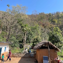 A rural setting with a thatched-roof hut and a solar panel in the foreground, surrounded by lush greenery and hills. A woman and a child are seen interacting, while another person stands near the hut, engaging in daily chores under a clear blue sky.