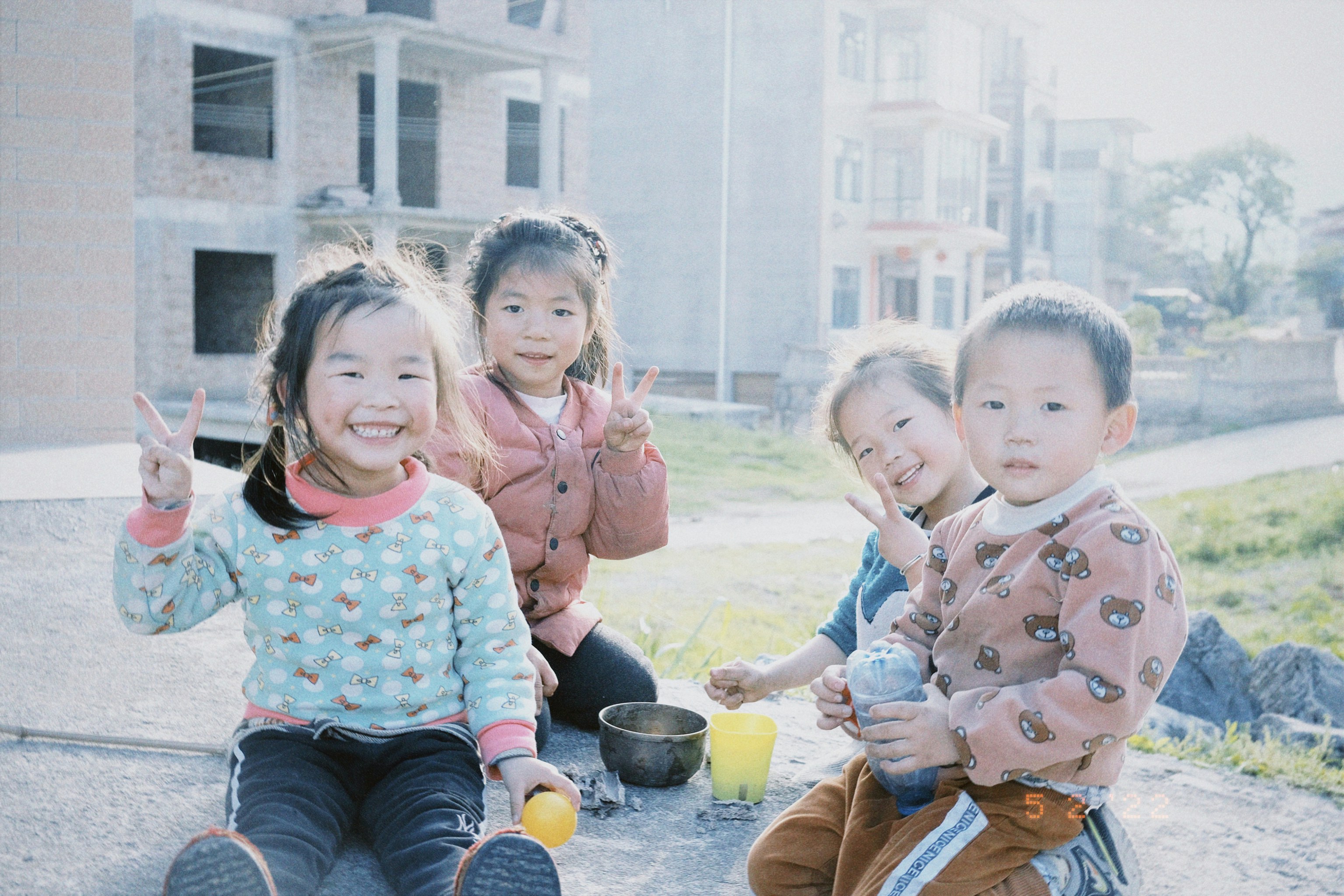 Four children are seated outdoors with an unfinished building in the background. They are smiling and making peace signs with their hands. The ground is made of concrete, and there are some toys and cups around them. The setting appears to be a grassy and open area with a bright and sunny atmosphere.