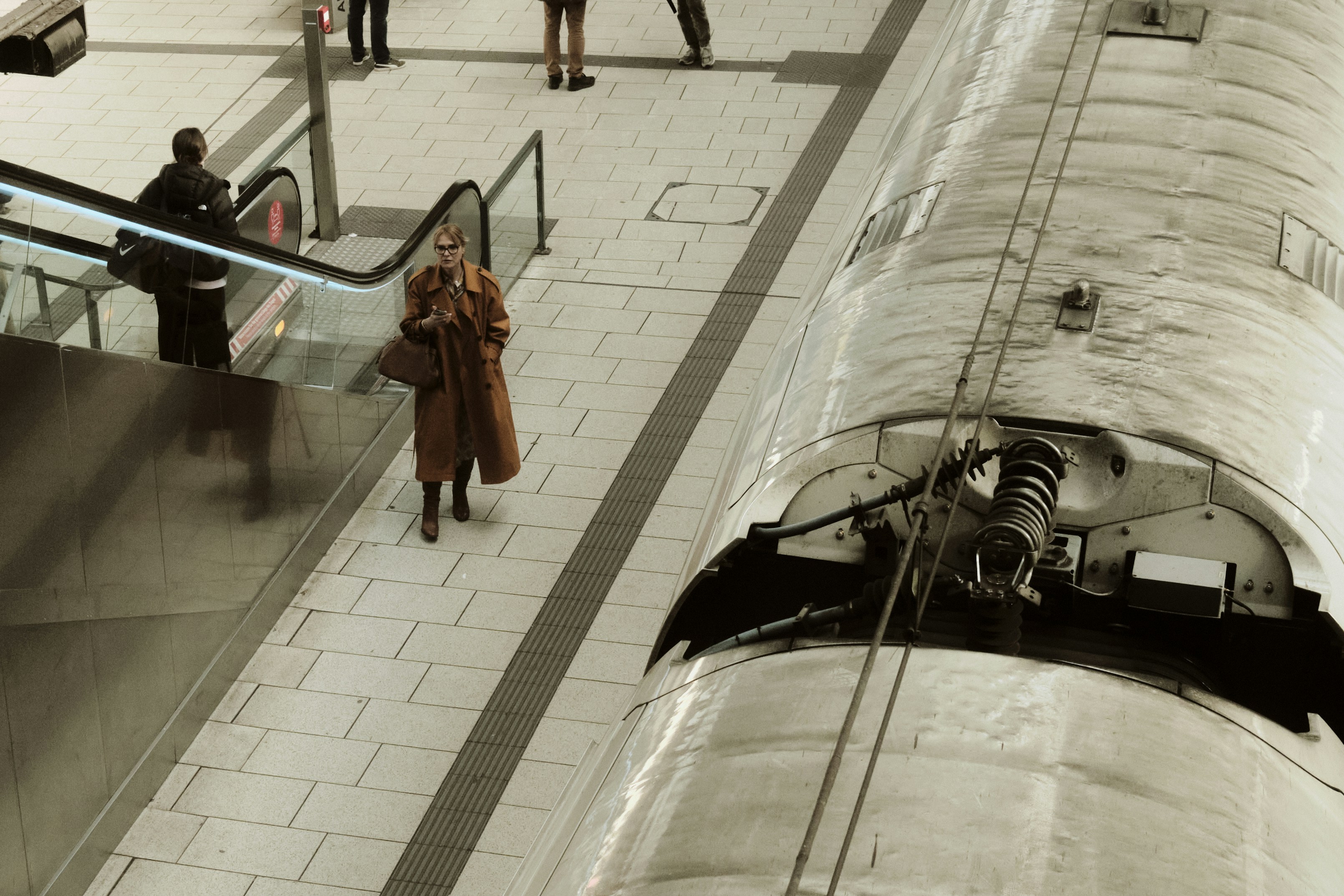 a woman in a trench coat walking down a walkway next to an escalator