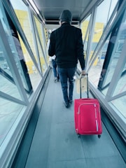 A person wearing a dark jacket and beanie walks down an airport jet bridge while pulling a red suitcase. The passageway has glass walls with metal frames, and another person is visible in the distance. The walls have a yellow pattern on them, and the ground outside is visible through the windows.