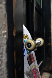 A skateboard with a worn appearance is leaning against a dark wooden wall. The deck features various stickers, including bright yellow smiley faces and abstract designs. The skateboard's wheels are off-white with signs of use.