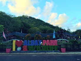 A colorful sign with large, bold letters saying 'I ❤️ ALBAY PARK & WILDLIFE' is situated at the forefront, surrounded by lush greenery. The background features a small hill covered with dense trees. The sky is bright with scattered fluffy clouds, adding a serene atmosphere.