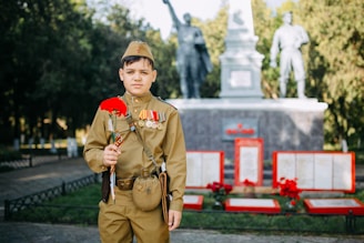A young boy dressed in a military uniform adorned with medals and ribbons stands in front of a monument. He holds a red carnation in his hand, while the background features statues and memorial plaques surrounded by greenery.