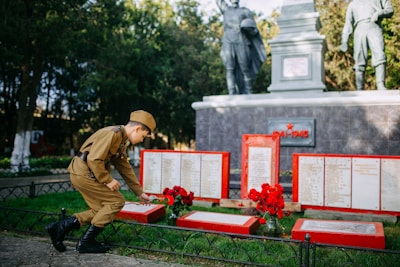 A young person in a military uniform is placing a flower on a memorial site. The memorial consists of plaques with inscriptions and bright red frames. In the background, there are statues of soldiers and trees.