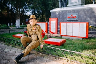 A young person dressed in military uniform sits on a low fence in a memorial park setting. Behind them are red-bordered plaques and a memorial honoring the years 1941-1945, suggesting a World War II monument. Bright greenery contrasts with the gray stone and red accents of the memorial.