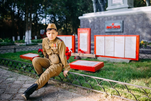A young person dressed in military uniform sits on a low fence in a memorial park setting. Behind them are red-bordered plaques and a memorial honoring the years 1941-1945, suggesting a World War II monument. Bright greenery contrasts with the gray stone and red accents of the memorial.