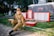 A young person dressed in military uniform sits on a low fence in a memorial park setting. Behind them are red-bordered plaques and a memorial honoring the years 1941-1945, suggesting a World War II monument. Bright greenery contrasts with the gray stone and red accents of the memorial.