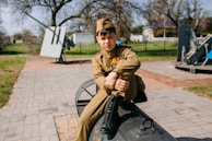 A young boy wearing a military-style uniform with medals sits on a large piece of military equipment, possibly an artillery gun, in an outdoor setting. The background features some paved pathways, grassy areas, and trees, along with additional military equipment. The boy appears pensive or thoughtful.