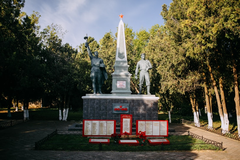 A war memorial featuring two statues of soldiers standing on either side of an obelisk, with one holding a flag and the other saluting. The base of the monument has plaques with writing, and red stars and flowers add to the commemorative feel. The setting is a park surrounded by tall trees.
