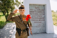 A young person in a military uniform salutes while holding red carnations in front of a stone monument with text commemorating a historical event. The setting is outdoors with greenery in the background.