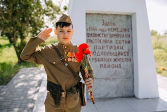 A young person in a military uniform salutes while holding red carnations in front of a stone monument with text commemorating a historical event. The setting is outdoors with greenery in the background.