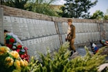 A child in a military-style uniform stands solemnly in front of a memorial wall inscribed with names and dates from the years 1941 to 1945. The wall is adorned with several floral wreaths, featuring a variety of vividly colored flowers. The background consists of lush greenery, suggesting a serene outdoor setting.