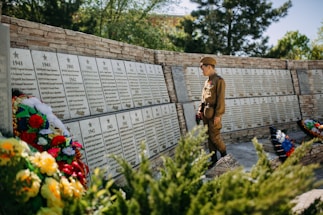 A child in a military-style uniform stands solemnly in front of a memorial wall inscribed with names and dates from the years 1941 to 1945. The wall is adorned with several floral wreaths, featuring a variety of vividly colored flowers. The background consists of lush greenery, suggesting a serene outdoor setting.