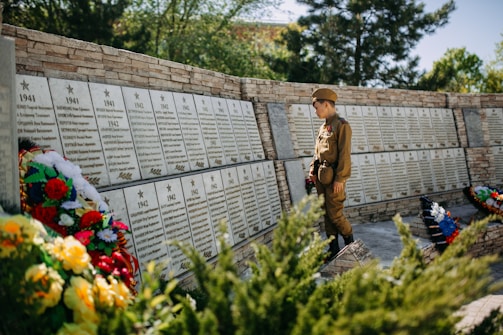 A child in a military-style uniform stands solemnly in front of a memorial wall inscribed with names and dates from the years 1941 to 1945. The wall is adorned with several floral wreaths, featuring a variety of vividly colored flowers. The background consists of lush greenery, suggesting a serene outdoor setting.