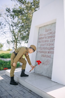 A person in a military uniform is placing red flowers at the base of a memorial stone with an inscription. The setting is outdoors, with trees and grass visible in the background. The flowers and the person's uniform contrast with the white and gray tones of the monument.