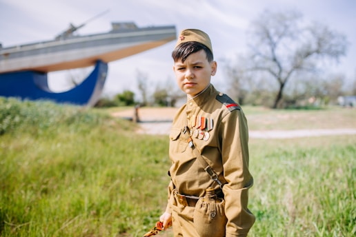 A young person in a military uniform stands in a grassy area with a large, elevated ship structure in the background. The uniform is adorned with several medals and ribbons. Leafless trees and a soft blue sky are visible in the background, suggesting a calm and open outdoor setting.