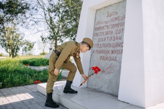 A person dressed in military-style clothing is laying flowers at a memorial with an inscription. The commemorative stone has red text and artwork, and the setting is outdoors with green grass and trees in the background.