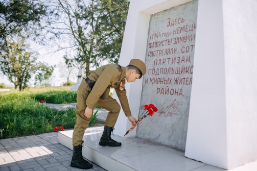 A person dressed in military-style clothing is laying flowers at a memorial with an inscription. The commemorative stone has red text and artwork, and the setting is outdoors with green grass and trees in the background.