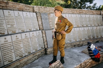 A young boy in a military uniform stands in front of a war memorial wall decorated with plaques listing names and dates. The boy is adorned with medals and insignia on his uniform and holds a respectful stance. There are flowers laid at the base of the memorial wall.
