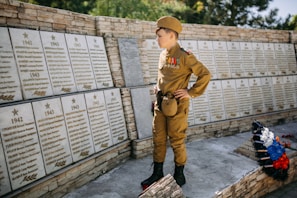 A young boy in a military uniform stands in front of a war memorial wall decorated with plaques listing names and dates. The boy is adorned with medals and insignia on his uniform and holds a respectful stance. There are flowers laid at the base of the memorial wall.