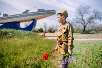 A young person dressed in a vintage military uniform holds red and purple flowers while standing on a grassy field. In the background, there is a large naval monument structured like a ship. The scene is bright with a clear sky and sparse trees.