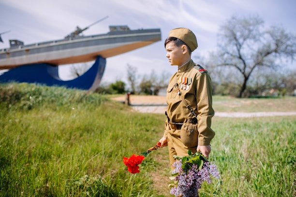 A young person dressed in a vintage military uniform holds red and purple flowers while standing on a grassy field. In the background, there is a large naval monument structured like a ship. The scene is bright with a clear sky and sparse trees.