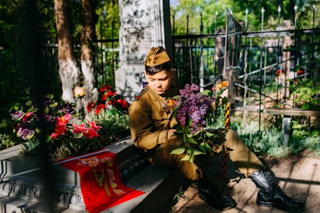 A young person wearing a military uniform sits beside a gravestone in a cemetery. They hold a bouquet of purple flowers and are surrounded by vibrant red and purple flowers. Sunlight filters through the trees, casting dappled shadows on the scene.