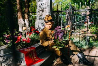 A young person wearing a military uniform sits beside a gravestone in a cemetery. They hold a bouquet of purple flowers and are surrounded by vibrant red and purple flowers. Sunlight filters through the trees, casting dappled shadows on the scene.
