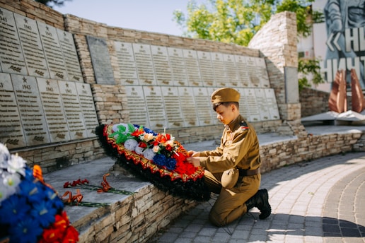 A young person in a military uniform kneels in front of a memorial wall adorned with plaques that likely list names and dates. They are thoughtfully arranging a wreath that is decorated with colorful flowers, including red, white, and blue hues. The backdrop consists of a stone wall with engraved plaques and additional floral arrangements on the ground. The setting is outdoors, and the lighting suggests a sunny day.