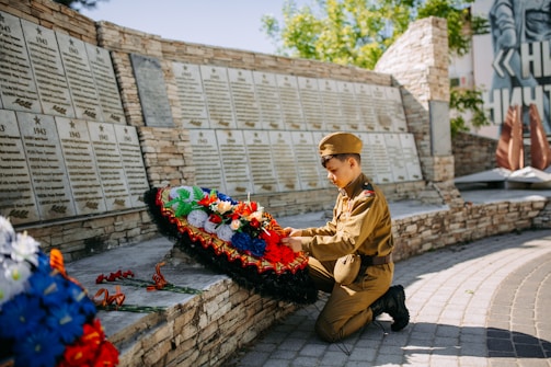 A young person in a military uniform kneels in front of a memorial wall adorned with plaques that likely list names and dates. They are thoughtfully arranging a wreath that is decorated with colorful flowers, including red, white, and blue hues. The backdrop consists of a stone wall with engraved plaques and additional floral arrangements on the ground. The setting is outdoors, and the lighting suggests a sunny day.