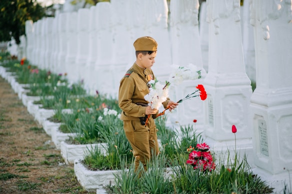 A young child in a military-style uniform is holding a bouquet of white and red flowers while standing in front of a row of white gravestones. The setting appears to be a cemetery with neatly arranged green plants and flowers around the gravestones.