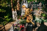 A person in military uniform tends to a grave decorated with colorful flowers. Sunlight filters through nearby trees, casting dappled shadows across the scene. The grave is situated in an old cemetery surrounded by iron fencing, with various headstones visible in the background.