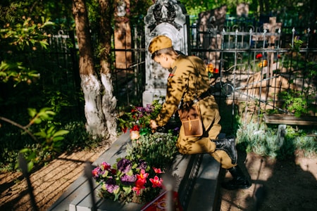 A person in military uniform tends to a grave decorated with colorful flowers. Sunlight filters through nearby trees, casting dappled shadows across the scene. The grave is situated in an old cemetery surrounded by iron fencing, with various headstones visible in the background.
