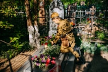 A person in military uniform tends to a grave decorated with colorful flowers. Sunlight filters through nearby trees, casting dappled shadows across the scene. The grave is situated in an old cemetery surrounded by iron fencing, with various headstones visible in the background.