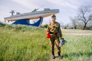 A young person stands in a grassy area wearing a military-style uniform adorned with medals. They hold a bouquet of red and purple flowers. In the background, a large ship sculpture is visible, set against a backdrop of trees and a blue sky.