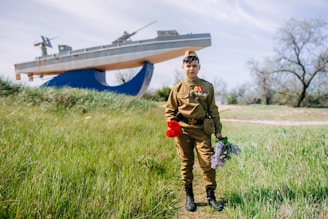 A young person stands in a grassy area wearing a military-style uniform adorned with medals. They hold a bouquet of red and purple flowers. In the background, a large ship sculpture is visible, set against a backdrop of trees and a blue sky.