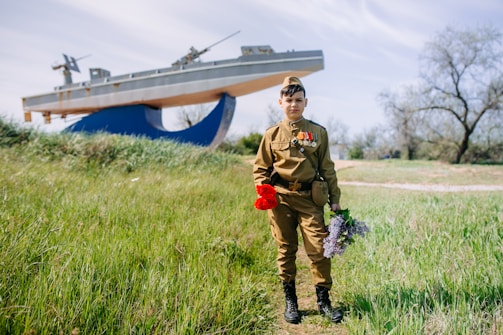 A young person stands in a grassy area wearing a military-style uniform adorned with medals. They hold a bouquet of red and purple flowers. In the background, a large ship sculpture is visible, set against a backdrop of trees and a blue sky.
