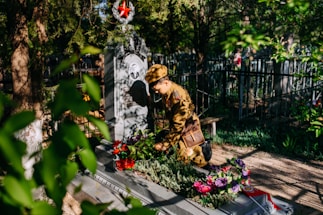 A person in a military uniform is kneeling beside a decorated grave in a cemetery, surrounded by greenery. There are colorful flowers placed on the grave and a prominent red star emblem on the headstone. Sunlight filters through the trees, casting shadows.