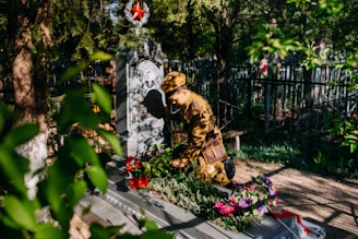 A person in a military uniform is kneeling beside a decorated grave in a cemetery, surrounded by greenery. There are colorful flowers placed on the grave and a prominent red star emblem on the headstone. Sunlight filters through the trees, casting shadows.