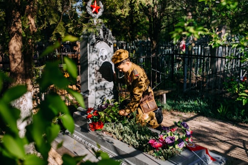 A person in a military uniform is kneeling beside a decorated grave in a cemetery, surrounded by greenery. There are colorful flowers placed on the grave and a prominent red star emblem on the headstone. Sunlight filters through the trees, casting shadows.