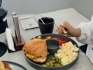 A plate of food featuring a croissant sandwich with cheese and meat sits alongside a serving of salad with lettuce and dressing, slices of tomato, pickles, and potato wedges. A person's hand with light green painted nails is resting near the plate on a light grey table, which also has a black cup with a straw, a napkin holder, and two metal cylinders.