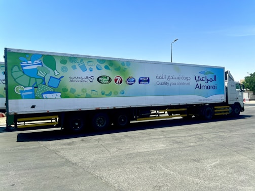 A large white truck with a trailer is parked on a road. The trailer features vibrant advertisements for a brand called Almarai, with the text 'Quality you can trust' written in both English and Arabic. The left side of the trailer displays illustrations of various food items, such as milk cartons, cheese, and limes.
