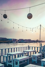 A serene waterfront view features a wooden deck with white benches and tables arranged in an outdoor seating area. Overhead, strings of lights and lanterns hang, contributing to a relaxed ambiance. In the background, a row of houses lines the water's edge under a softly lit sky, suggesting a peaceful evening setting.