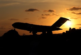 A silhouette of a large airplane is seen against the backdrop of a vibrant, orange sunset. The sky is dotted with scattered clouds, and the sun is seen near the horizon, casting an ethereal glow. There are silhouettes of people in the foreground, suggesting they are watching the plane.