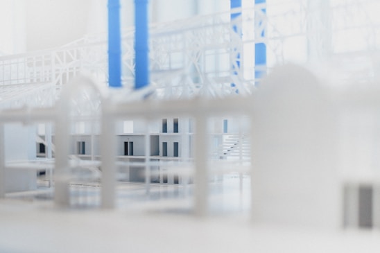 A close-up view of a scale architectural model featuring intricate white structures with geometric patterns. Vertical blue columns accentuate the composition, providing contrast against the white elements. The model has a modern and simplistic design.
