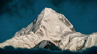 A majestic snow-covered mountain peak rises sharply against a deep blue sky. The light dusting of snow accentuates the rugged details of the mountain's surface.
