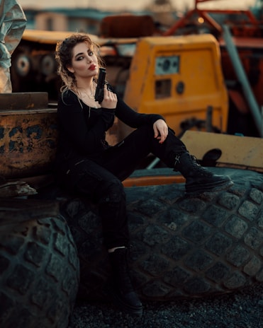 A person is sitting on large industrial tires, holding a handgun. They have curly hair, a confident expression, and are dressed in dark clothing. Surrounding them is an industrial setting with heavy machinery and worn textures, suggesting a gritty, urban environment.